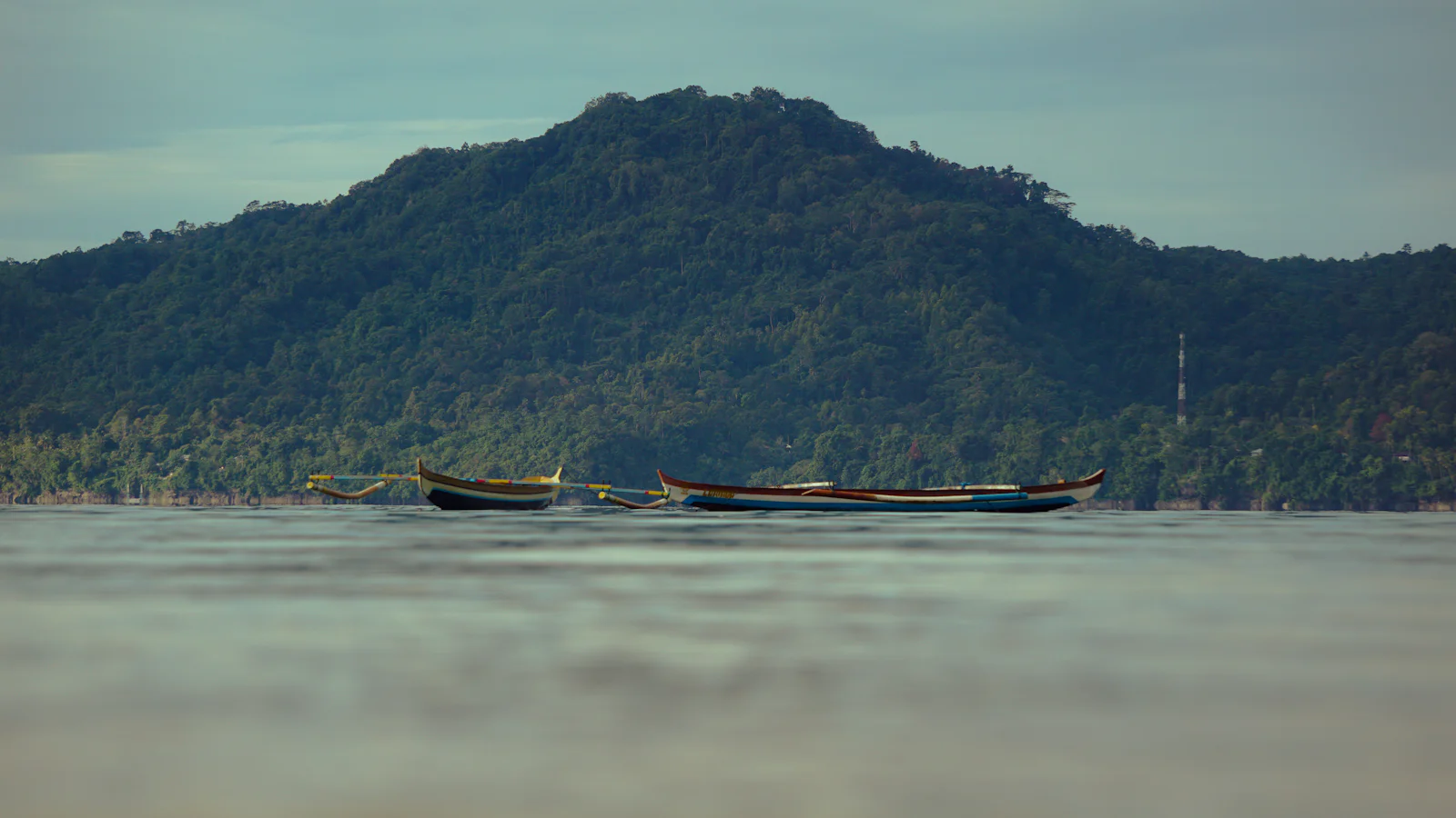 Two long boats on glassy water below a forested hill